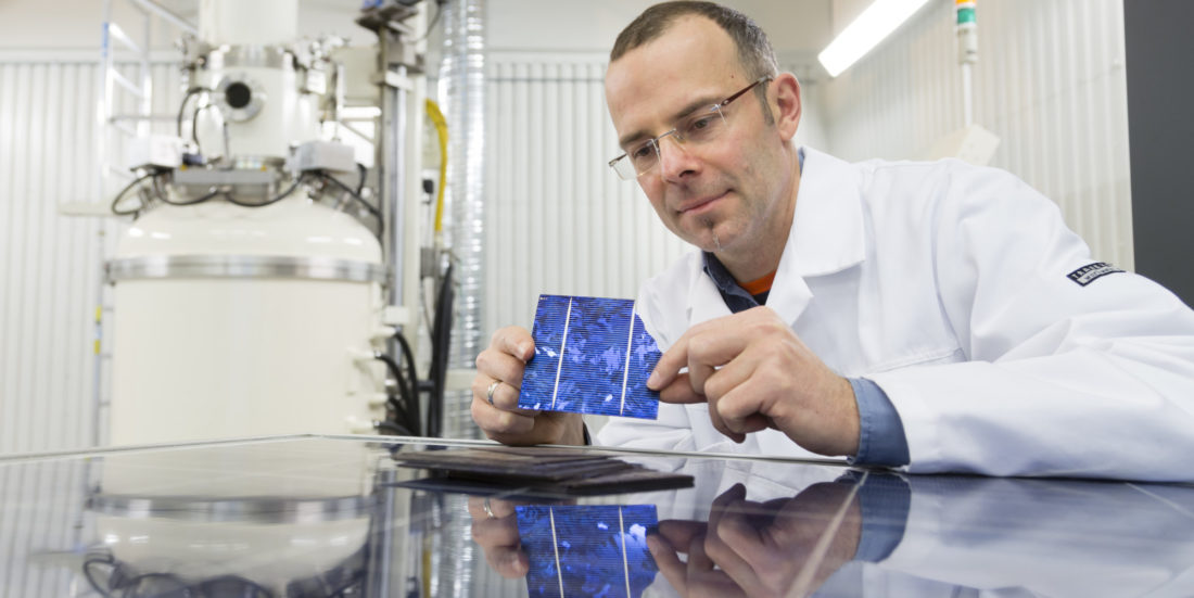 Mann i hvit frakk sitter på laboratorium der han ser på solcelle han holder i hendene.
