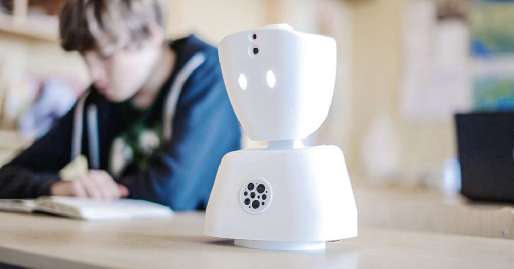 A small all-white robot stands on a school-desk while a kid studies in the background