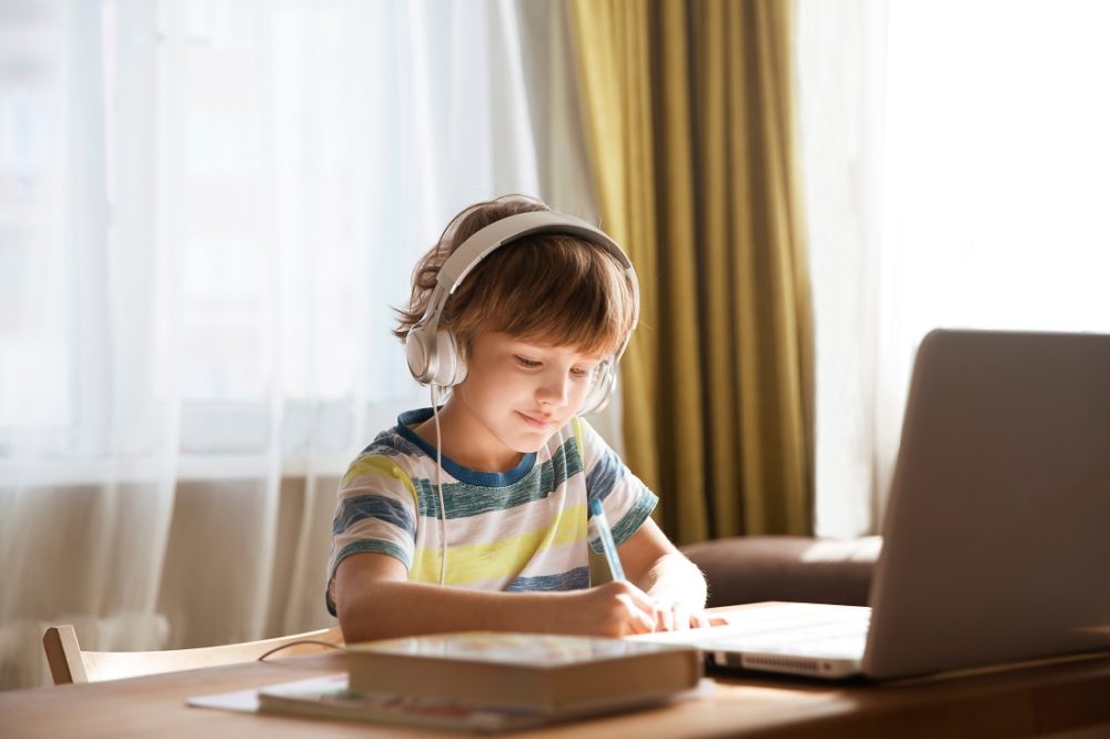 A child writing in a book with a computer in front of him and wearing a headset