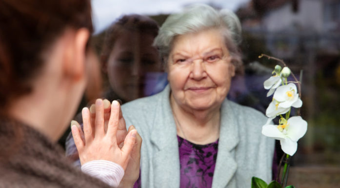 A young man greetings an old woman through a glass window. Both holding their hands on the window from opposite sides