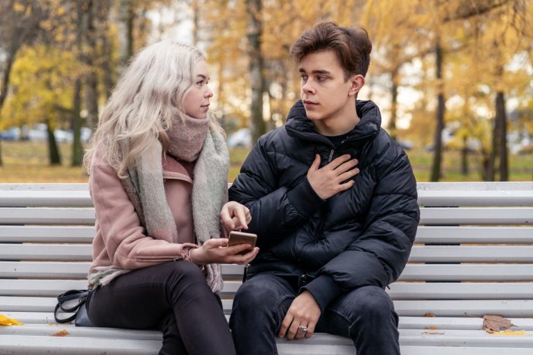 A man and woman on a bench. The woman points to something on her phone and the man acts innocent.