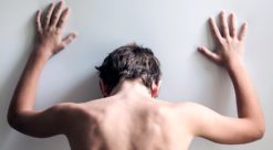 Rear view on shirtless back of young male teen with hands on white wall and head down in despair or desperation