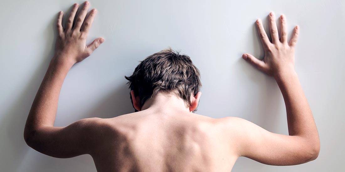 Rear view on shirtless back of young male teen with hands on white wall and head down in despair or desperation