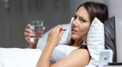 Woman in bed scowling at camera as she takes a pill. Photo.