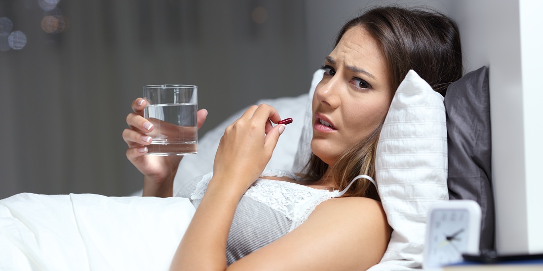 Woman in bed scowling at camera as she takes a pill. Photo.