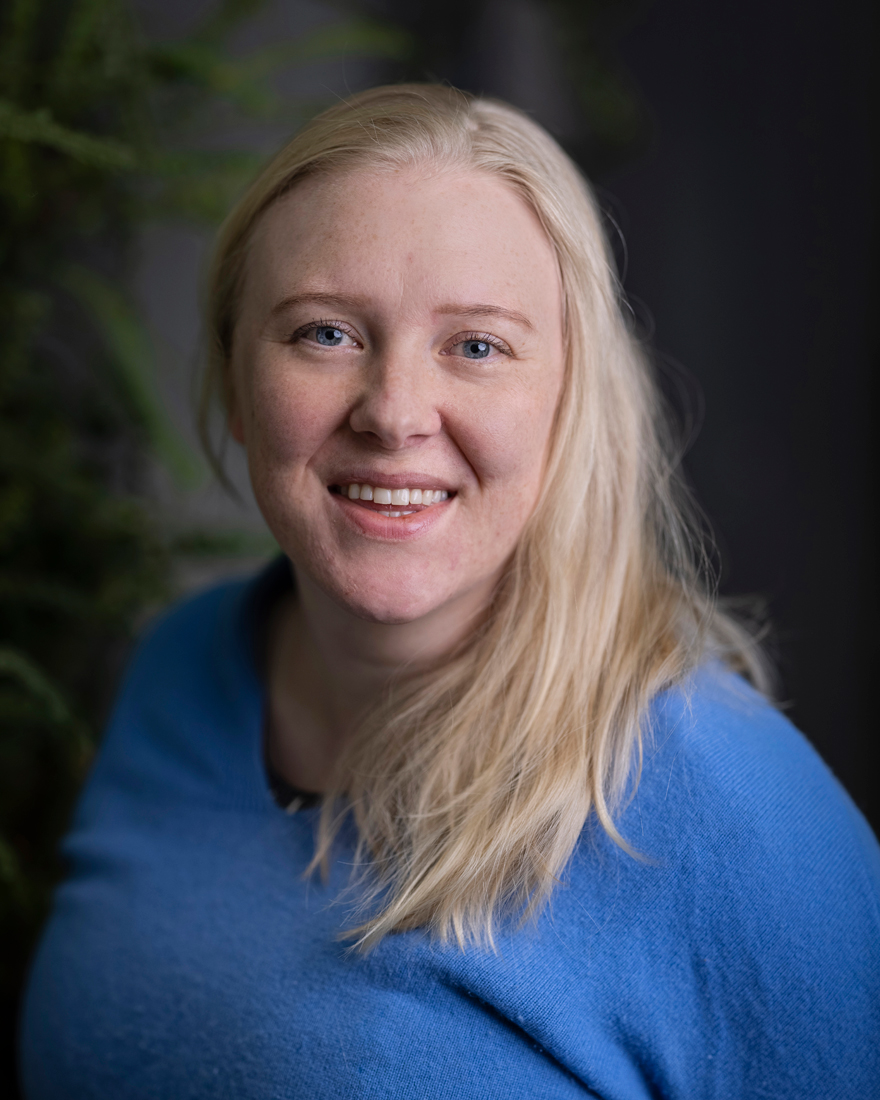 Smiling female alongside blonde tress and blue sweater. Photo.