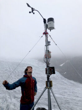A man standing by a weather station on a glacier in the fog.